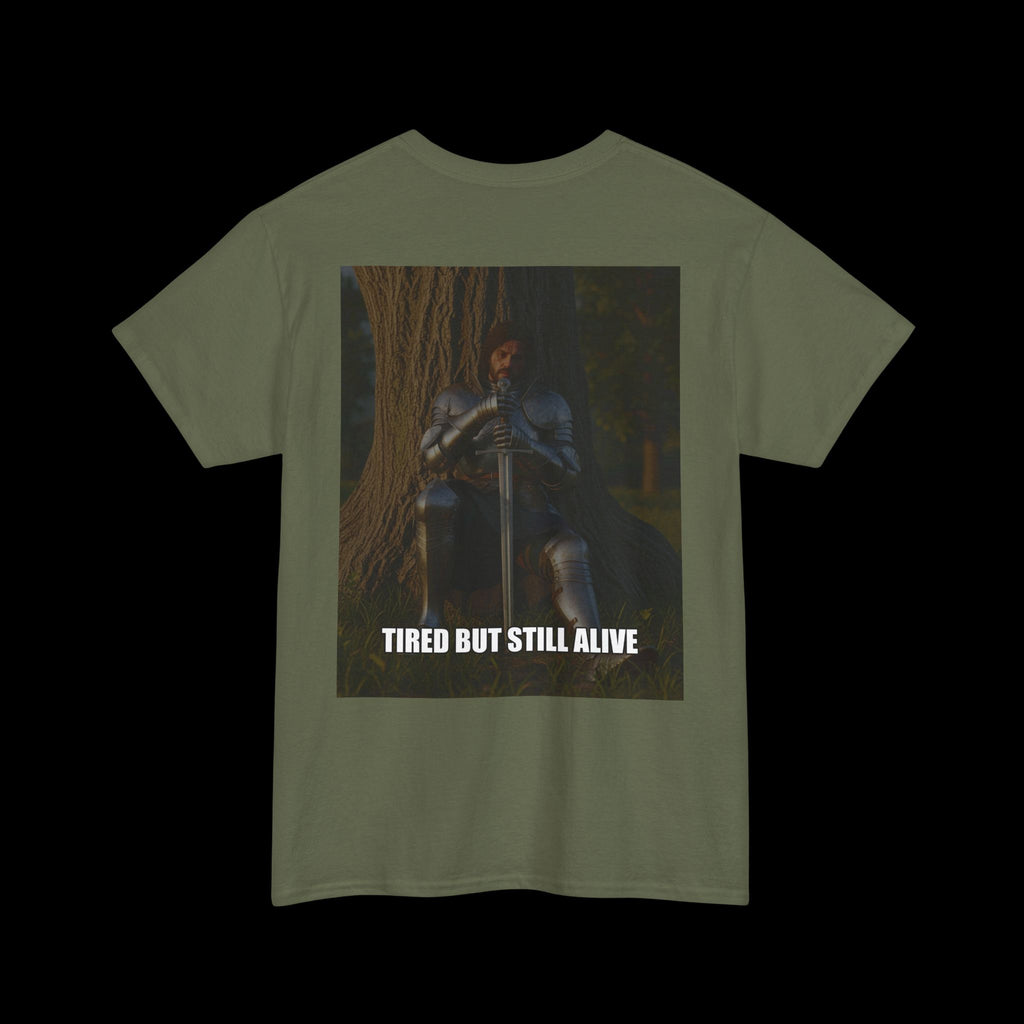 Olive green t-shirt featuring a graphic of a tired but resolute knight in armor sitting with a sword, and the text "TIRED BUT STILL ALIVE". Conveys a sense of endurance and quiet strength in an earthy tone.
